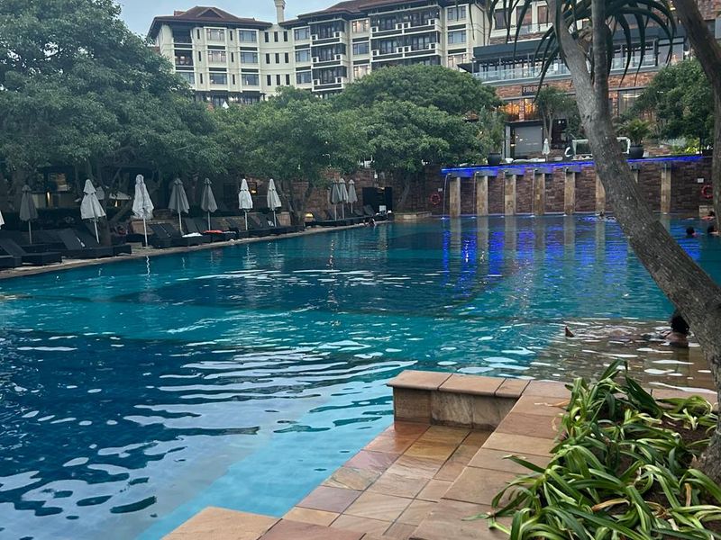 A well-maintained swimming pool area featuring stone tiling, a water feature, and landscaped surroundings within a residential complex.