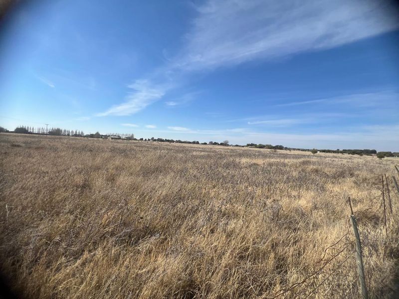 A wide-angle view of a large, flat, undeveloped residential plot featuring dry grass and perimeter wire fencing under a clear blue sky.