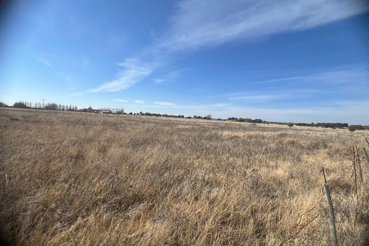 A wide-angle view of a large, flat, undeveloped residential plot featuring dry grass and perimeter wire fencing under a clear blue sky.