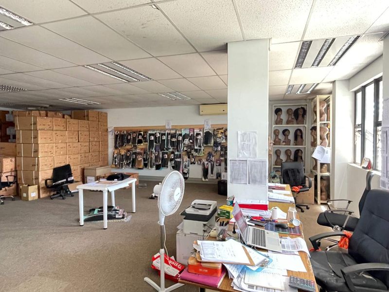 A commercial office space featuring a suspended ceiling, recessed lighting, wall-mounted air conditioning, and large windows, currently utilized for storage and administrative work.