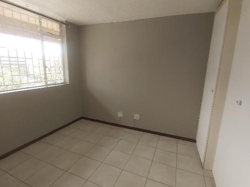 A bright, empty bedroom featuring neutral tiled flooring, a built-in cupboard, and security bars on the window.