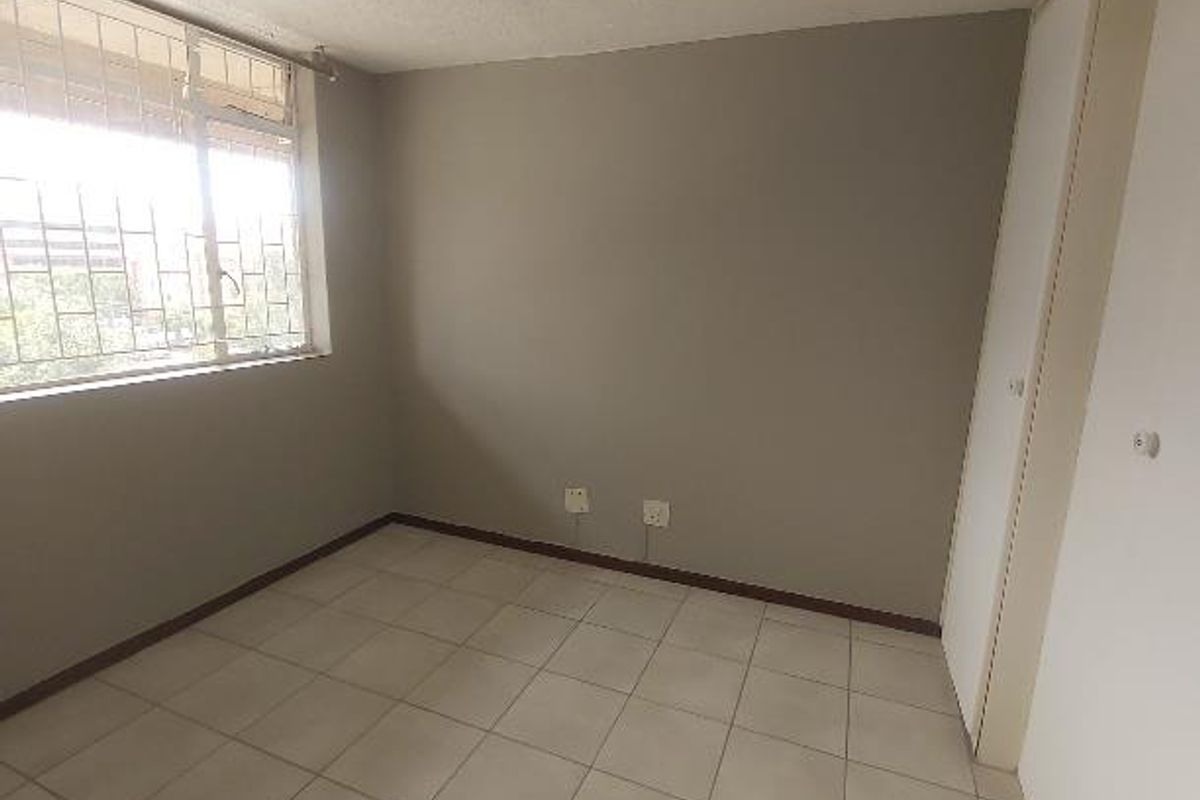 A bright, empty bedroom featuring neutral tiled flooring, a built-in cupboard, and security bars on the window.