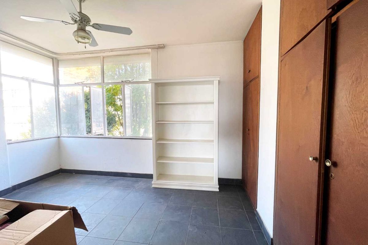 A bedroom featuring large windows, built-in wooden cupboards, and a ceiling fan, finished with dark floor tiles.