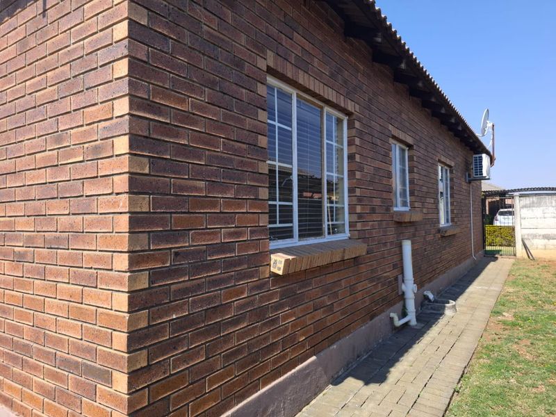 A side view of the townhouse featuring a durable face brick exterior, paved walkway, and functional external infrastructure including air conditioning and plumbing.