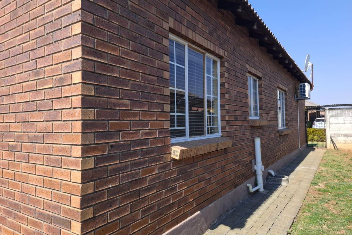A side view of the townhouse featuring a durable face brick exterior, paved walkway, and functional external infrastructure including air conditioning and plumbing.