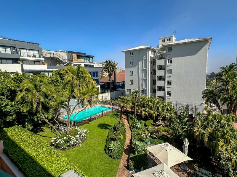 Elevated view of a well-maintained apartment complex garden featuring a swimming pool, lush landscaping, and paved pathways.