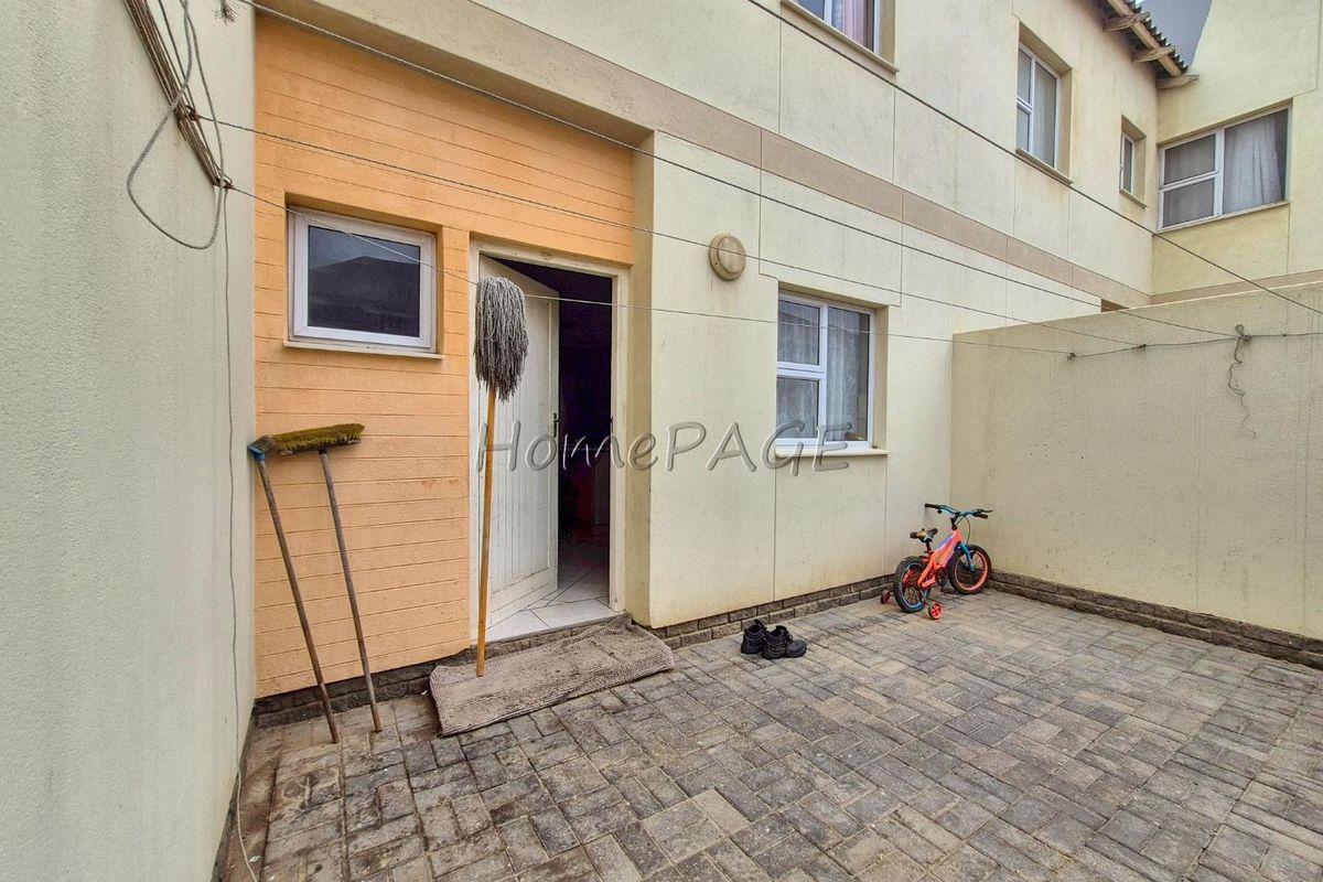 A paved rear courtyard area of a townhouse featuring exterior wall access, brick paving, and wall-mounted utility fixtures.