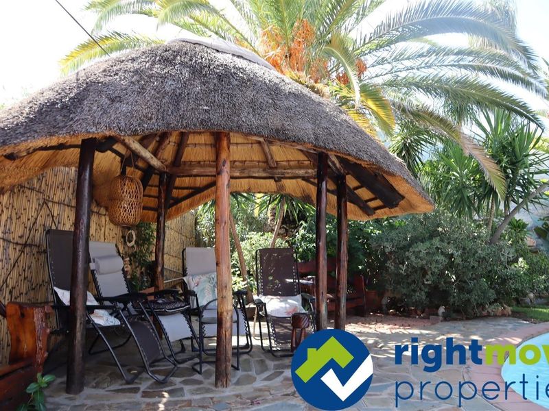 A rustic outdoor thatched lapa area featuring timber support posts and stone paving, providing a shaded retreat within the garden setting.