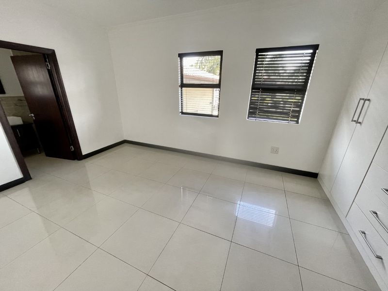 A bright, empty bedroom featuring neutral tiled flooring, built-in cupboards, and dark-framed windows with fitted venetian blinds.