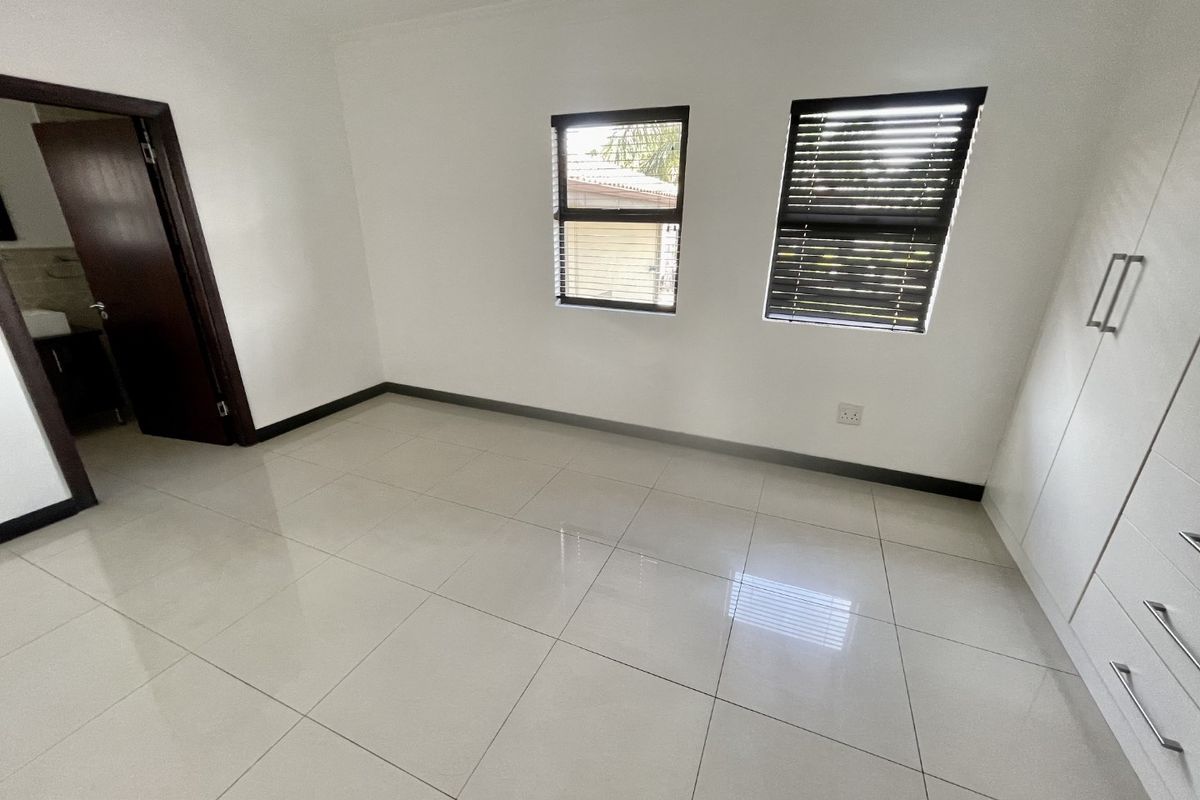 A bright, empty bedroom featuring neutral tiled flooring, built-in cupboards, and dark-framed windows with fitted venetian blinds.