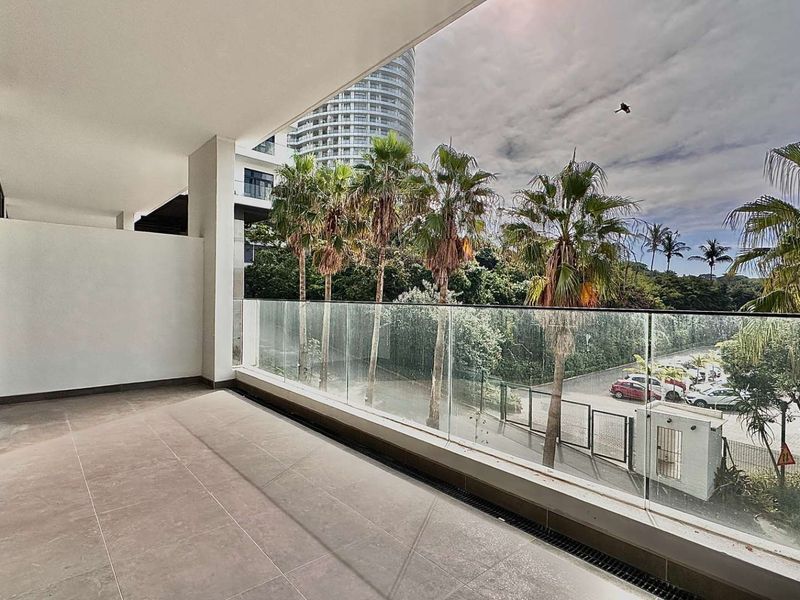 A spacious covered balcony featuring modern tiled flooring and a sleek glass balustrade, offering an open view of the surrounding area.