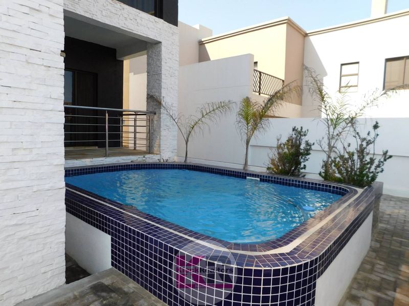 A compact, modern swimming pool area featuring blue mosaic tiling, stone-clad exterior walls, and a stainless steel balustrade leading to the patio.