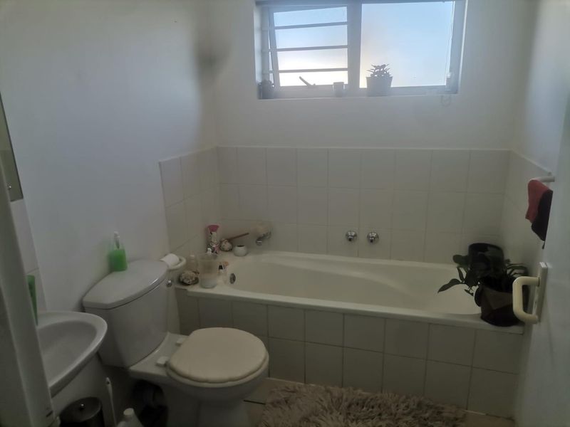 A functional bathroom featuring a built-in bathtub, white ceramic wall tiling, a standard toilet, and a pedestal basin with a louvered window.