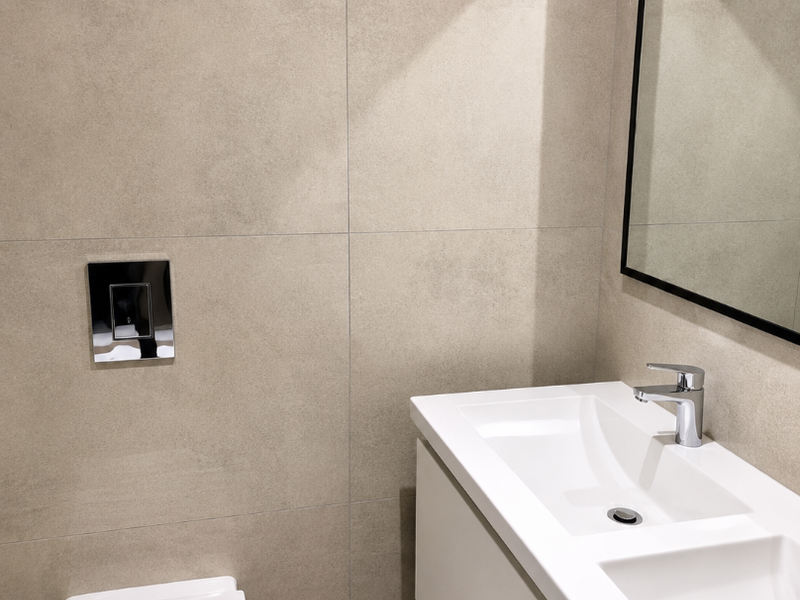 A modern, minimalist bathroom featuring a wall-mounted toilet, double vanity basin with chrome fixtures, and neutral large-format wall and floor tiles.
