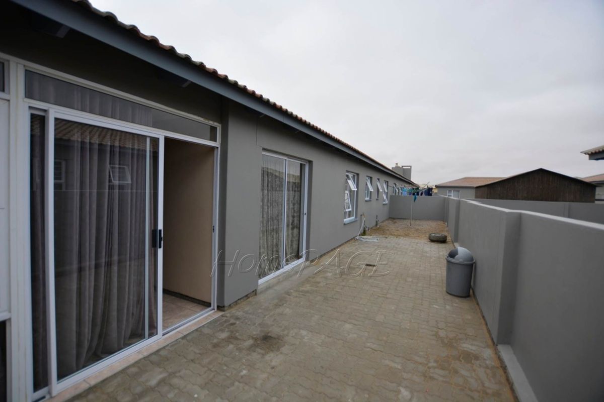 A rear exterior view of the townhouse featuring a paved courtyard, sliding glass doors, and a perimeter boundary wall.