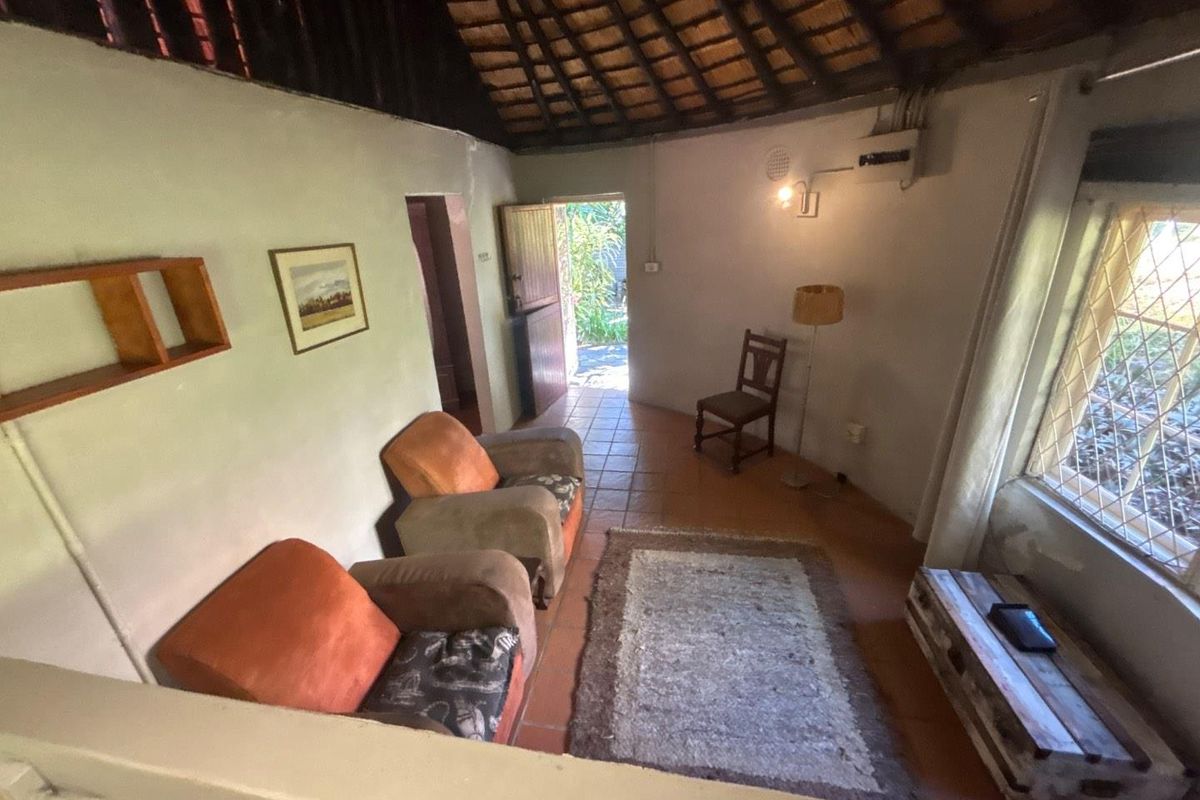 A cozy living room in a cottage featuring a traditional thatched roof with exposed rafters, terracotta tiled flooring, and diamond-pattern security window bars.