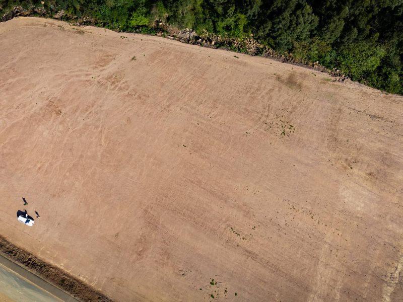Aerial view of a large, cleared industrial plot of land adjacent to an asphalt road, bordered by mature trees.