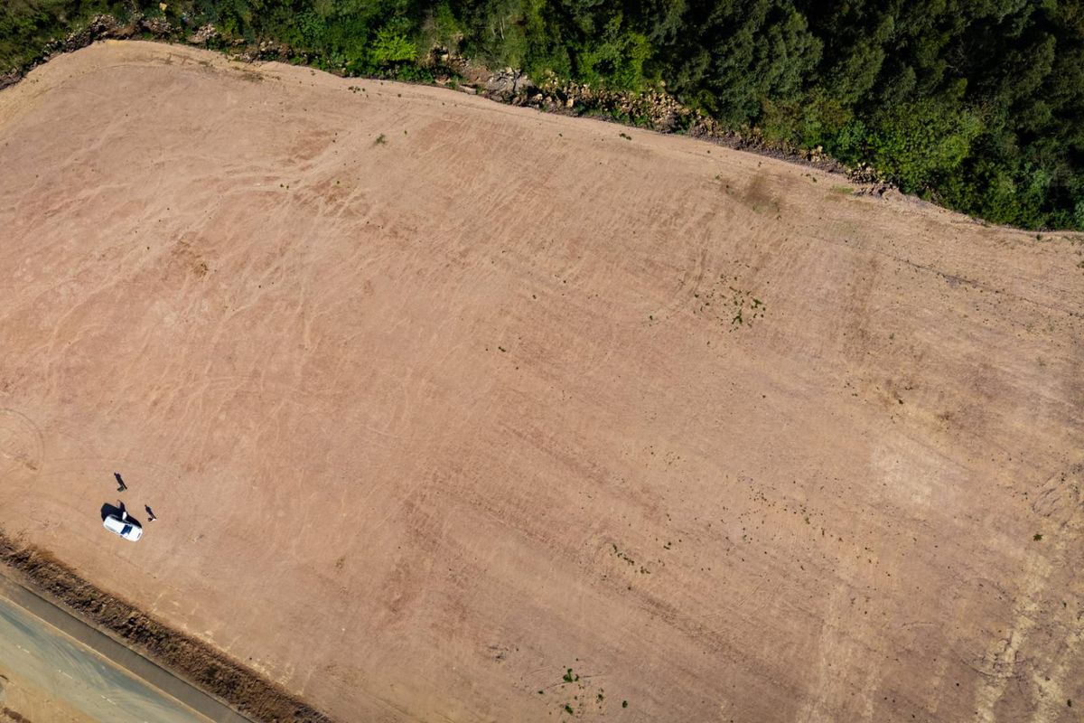 Aerial view of a large, cleared industrial plot of land adjacent to an asphalt road, bordered by mature trees.