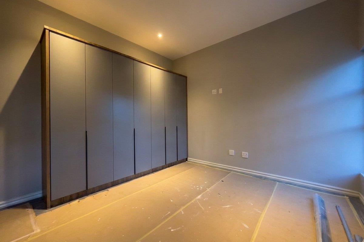A clean, empty bedroom featuring modern built-in cupboards with a sleek finish and recessed downlighting, currently undergoing final finishing touches.