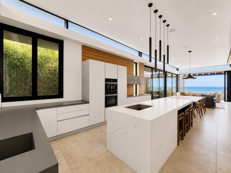Modern open-plan kitchen featuring a large stone island, built-in appliances, and expansive sliding doors opening to a scenic coastal view.