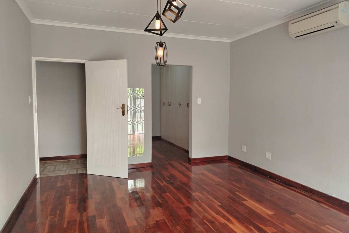 A spacious bedroom featuring polished hardwood flooring, neutral grey walls, a wall-mounted air conditioning unit, and built-in cupboard storage.