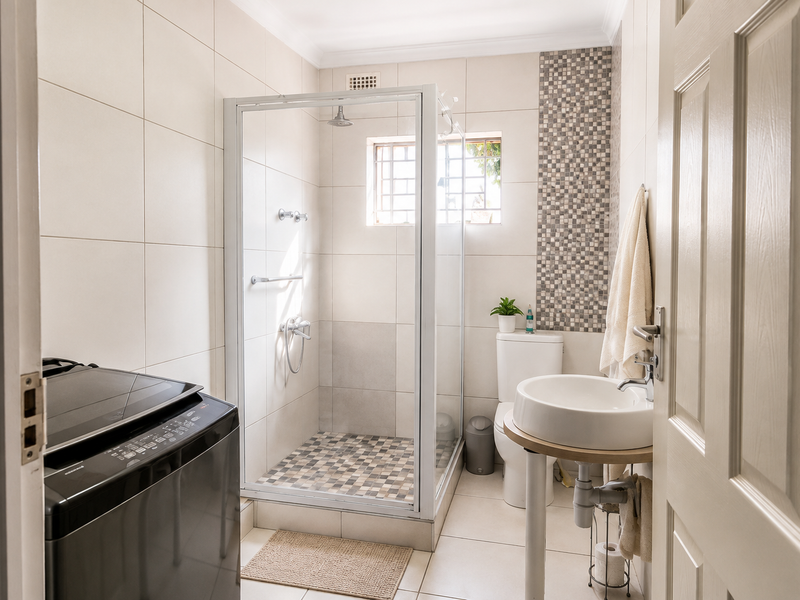 A clean, modern bathroom featuring a glass shower enclosure, mosaic tile accents, a pedestal sink, and a toilet, with neutral wall and floor tiling.