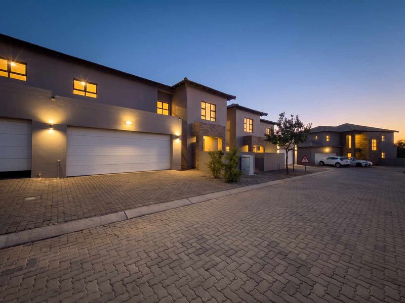 A modern townhouse exterior at dusk, featuring a paved driveway, double garage, and stone-clad architectural details with warm exterior lighting.