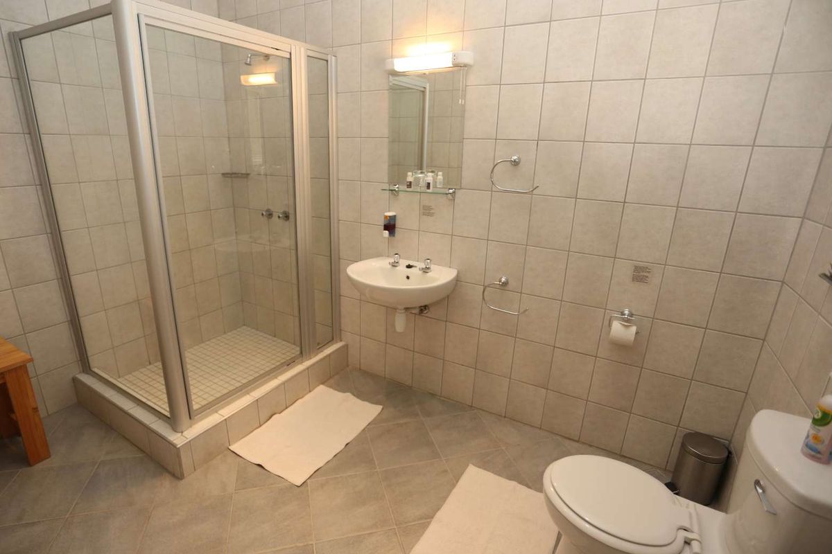 A functional hotel bathroom featuring a glass-enclosed shower, wall-mounted basin, and standard toilet with neutral ceramic wall and floor tiling.