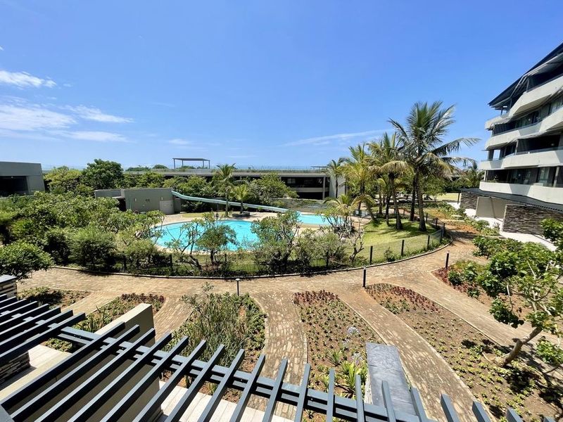 Elevated view of a well-maintained communal pool area featuring a water slide, landscaped gardens, and paved pathways within a modern apartment complex.