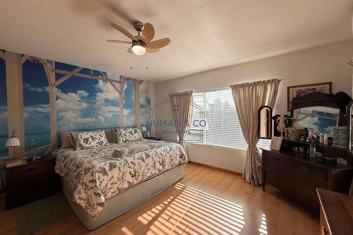 A well-lit bedroom featuring wood-look flooring, a ceiling fan, and a large window with blinds, providing a comfortable and functional living space.
