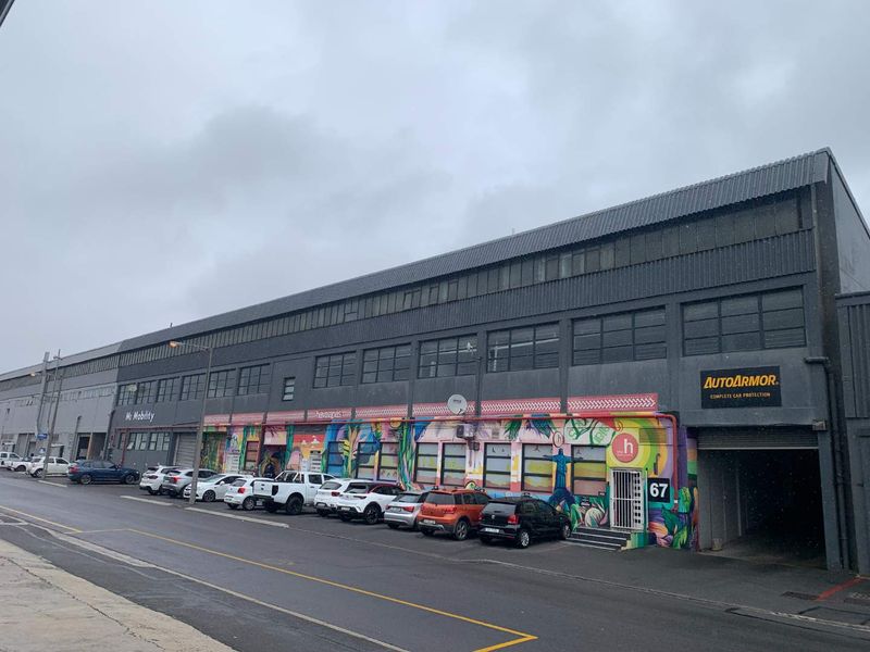 Street-level view of a multi-tenant industrial building featuring metal cladding, large multi-pane windows, and roller shutter access points.