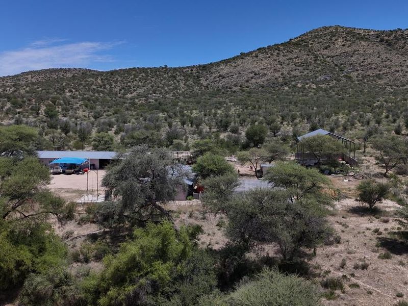 Aerial view of a rural farm property featuring multiple outbuildings, a carport, and expansive natural landscape set against a mountain backdrop.