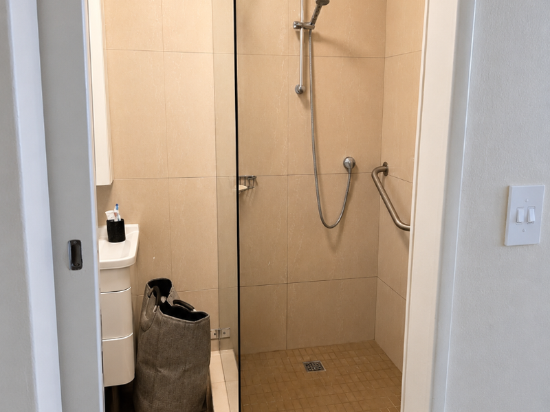 A compact bathroom featuring a tiled shower enclosure with a glass door, handheld shower head, grab rail, and a wall-mounted vanity basin.