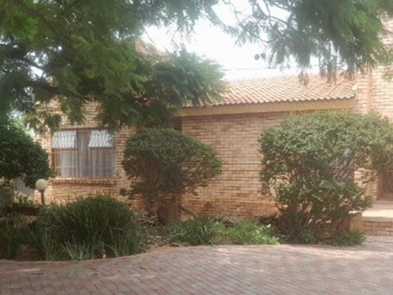 Front view of a face brick house featuring a tiled roof, security bars on windows, and a paved driveway surrounded by established garden greenery.