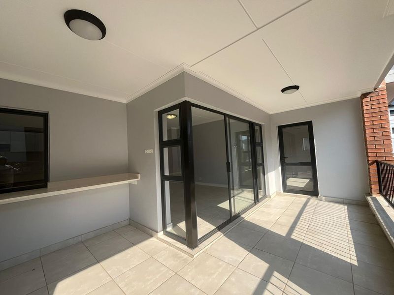 A clean, covered balcony featuring tiled flooring, a fixed breakfast bar, and modern black-framed glass doors leading into the apartment interior.