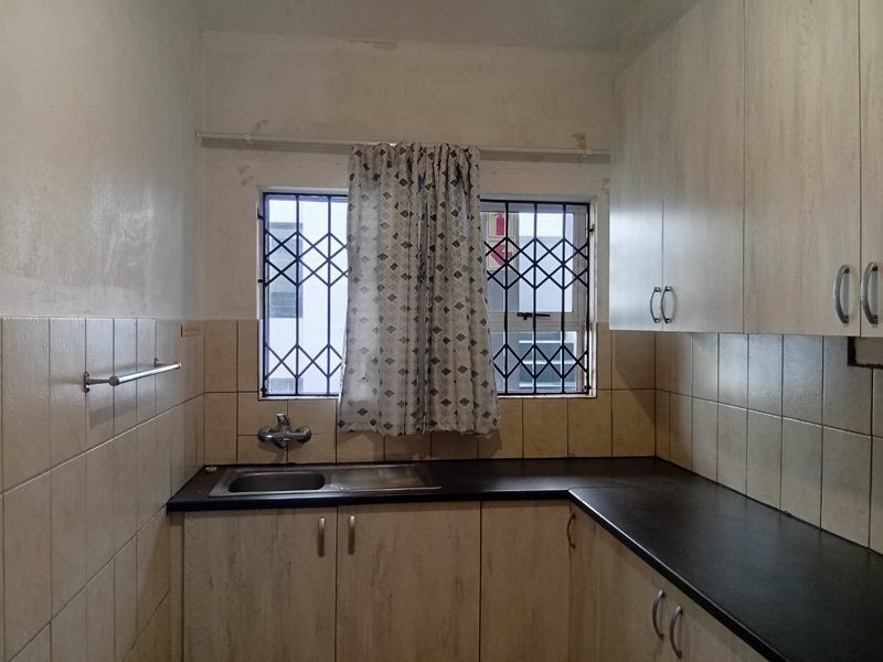 A compact kitchen featuring built-in cabinetry, dark laminate countertops, a stainless steel sink, and tiled splashbacks, with security bars installed on the windows.