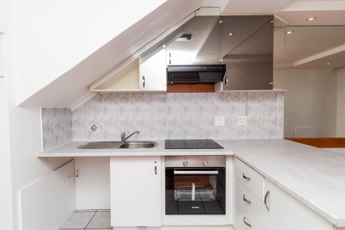 Compact kitchen featuring built-in oven, electric hob, extractor fan, stainless steel sink, and ample cabinetry with neutral tiled backsplash.