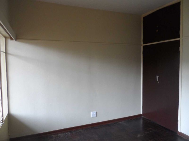 An empty bedroom featuring dark built-in cupboards, parquet flooring, and a large window providing natural light.