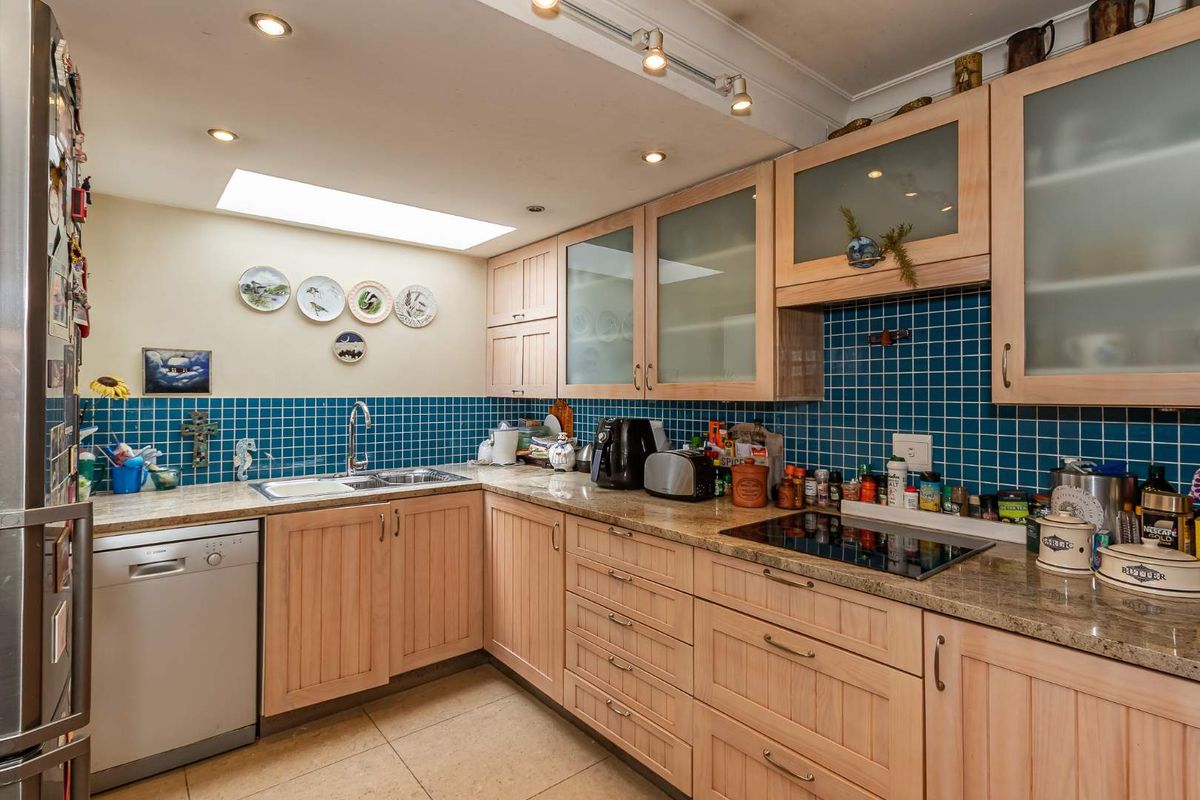 A functional kitchen featuring light wood cabinetry, granite countertops, blue tiled splashbacks, integrated hob, and a skylight providing natural illumination.