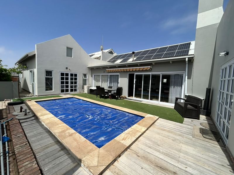 Rear outdoor entertainment area featuring a swimming pool with cover, timber decking, artificial lawn, and solar panels installed on the roof.
