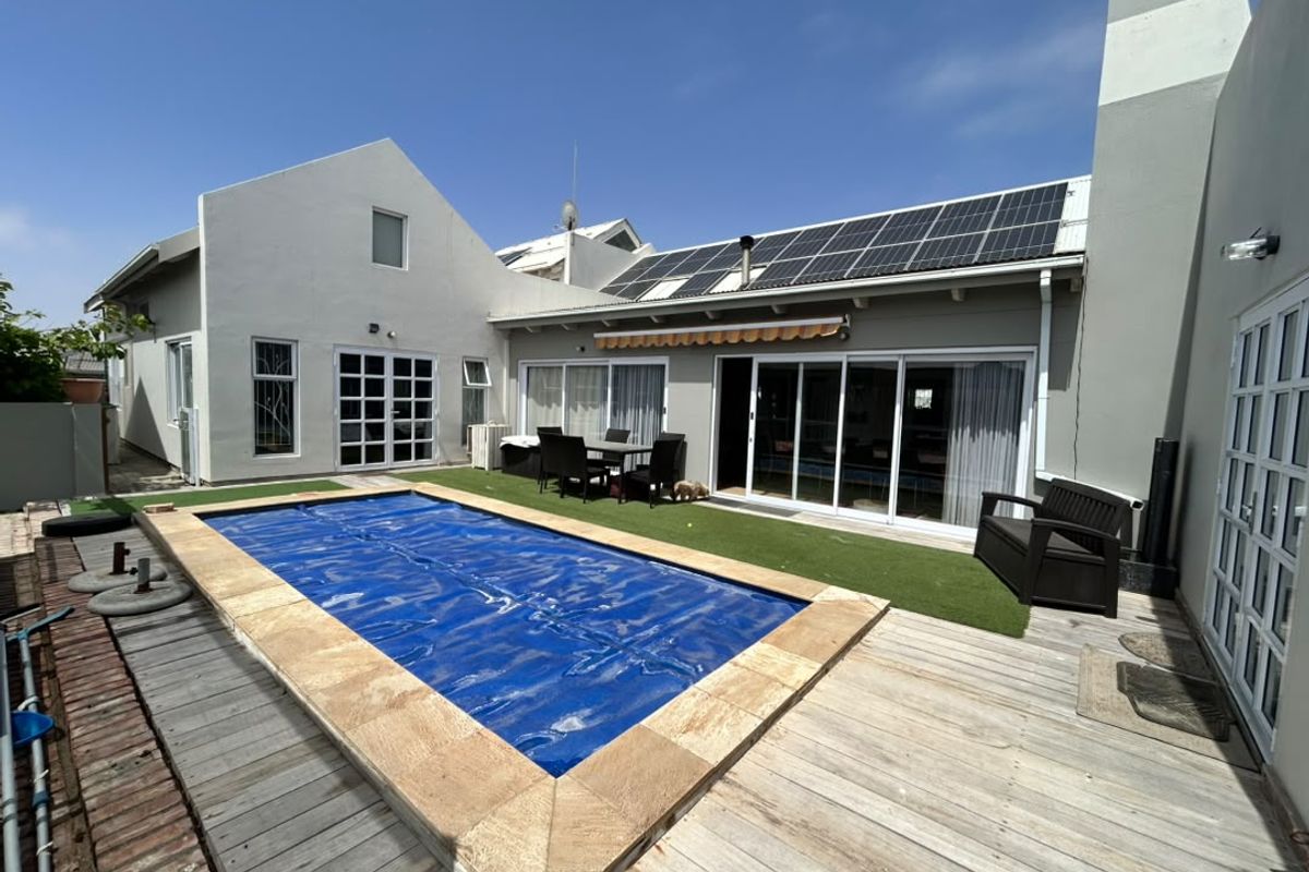 Rear outdoor entertainment area featuring a swimming pool with cover, timber decking, artificial lawn, and solar panels installed on the roof.