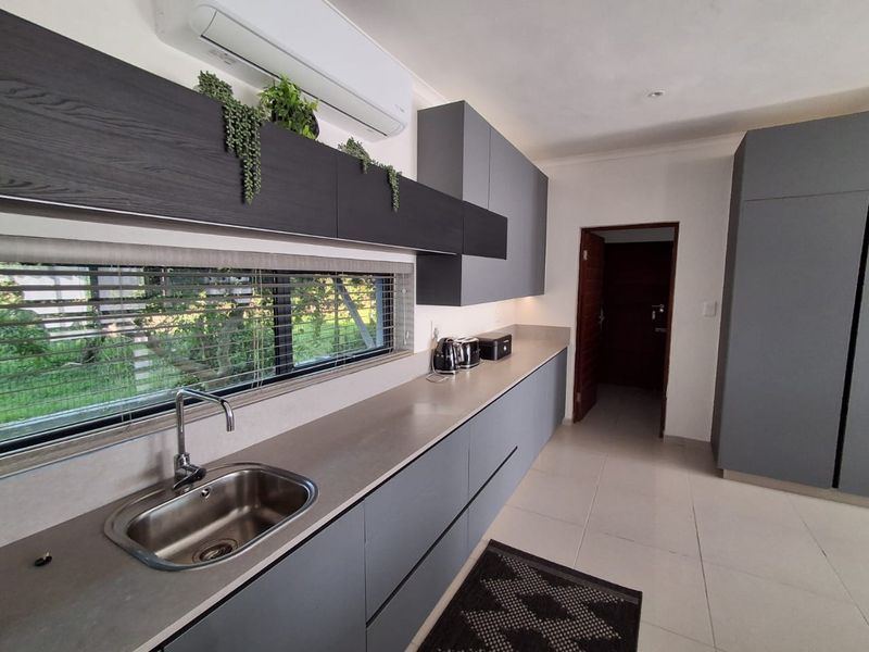 Modern kitchen featuring sleek grey cabinetry, stone countertops, stainless steel sink, and integrated air conditioning unit with large window overlooking the garden.