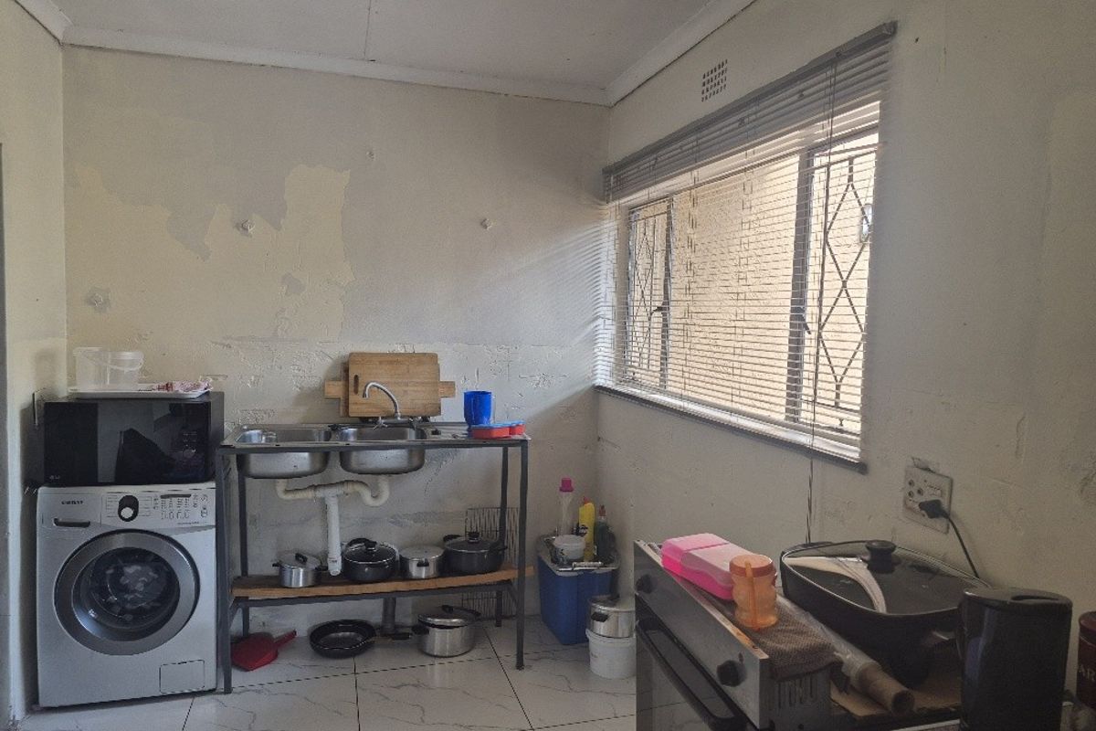 A functional kitchen area featuring tiled flooring, a double stainless steel sink, and a window with security bars, currently requiring wall surface repairs.