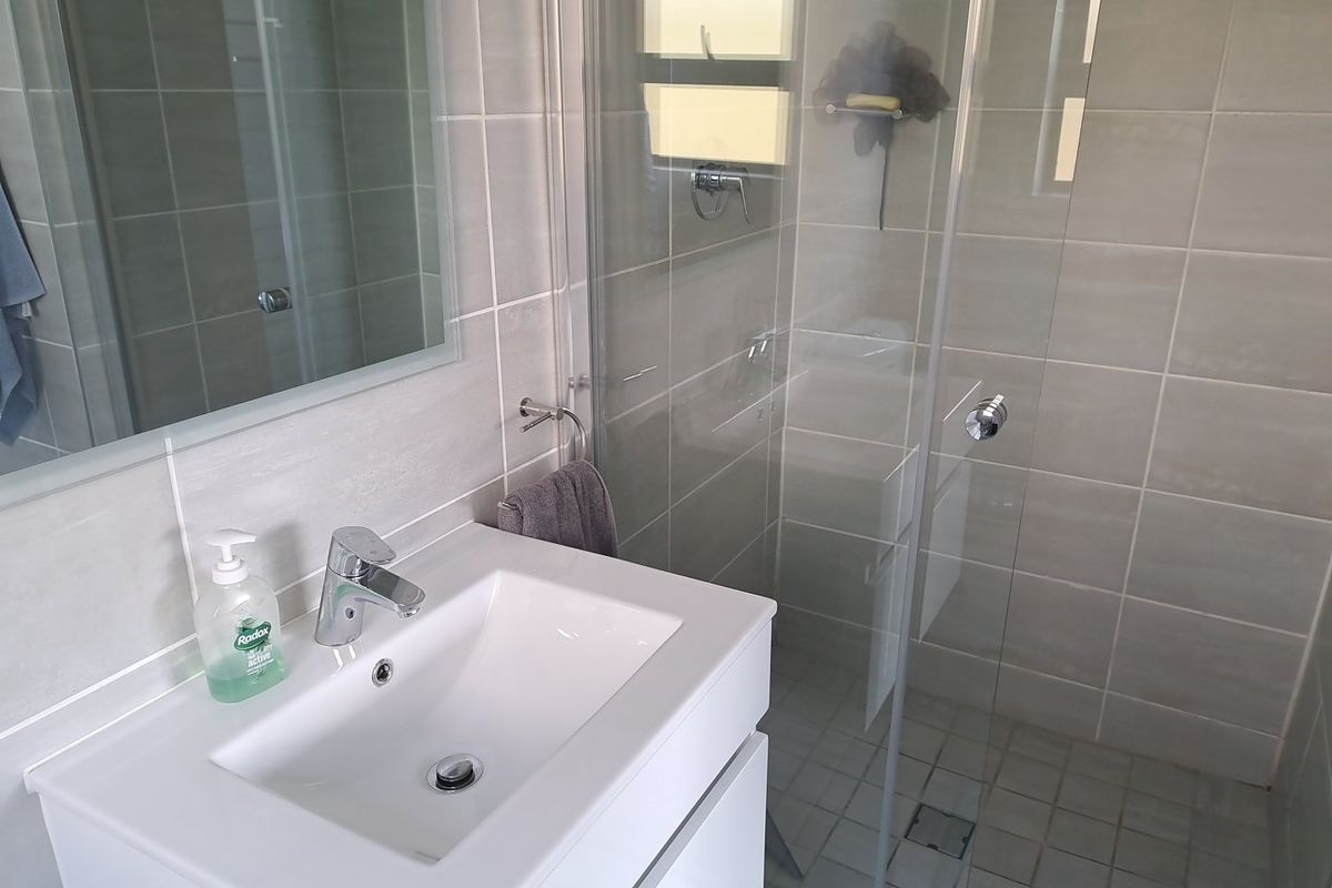 A modern bathroom featuring a white vanity with basin, wall-mounted mirror, and a glass-enclosed shower with neutral wall and floor tiling.