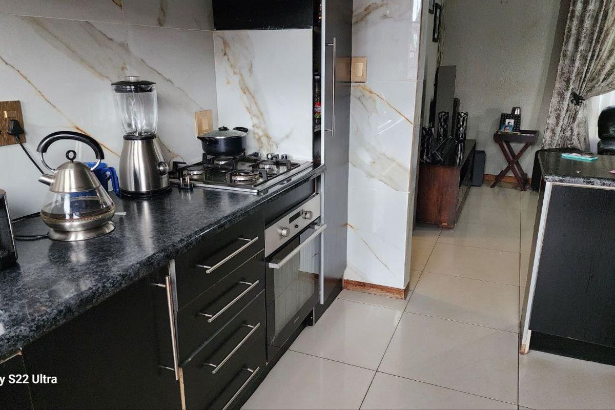Modern kitchen featuring dark cabinetry, granite countertops, built-in oven, gas hob, and marble-effect wall tiling, seamlessly flowing into the adjacent living area.