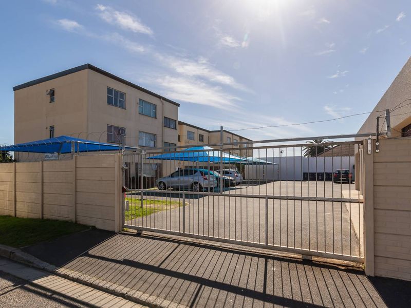 Exterior view of the apartment complex entrance featuring a secure gated driveway, asphalt parking area, and shade cloth carports.