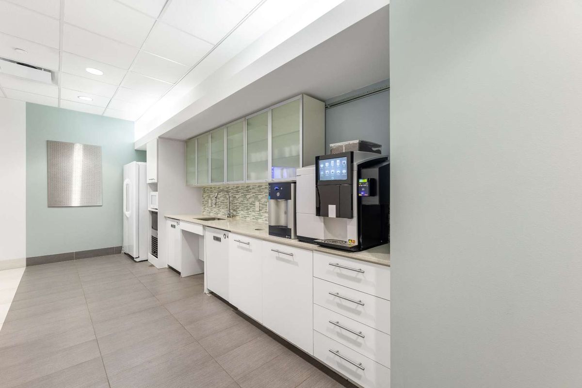 A modern office kitchenette featuring white cabinetry, stone countertops, a mosaic tile backsplash, and integrated sink with professional coffee station.