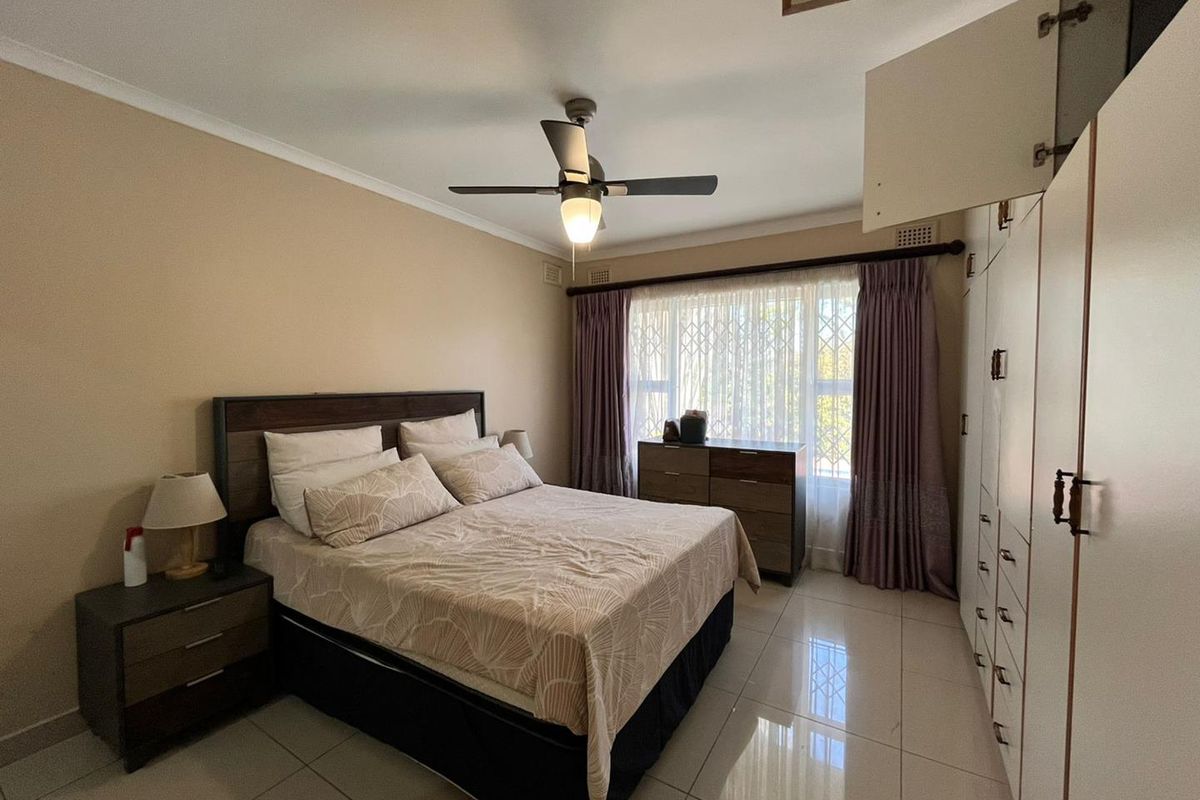 A well-lit bedroom featuring tiled flooring, a ceiling fan, and extensive built-in cupboard storage with a security gate on the window.