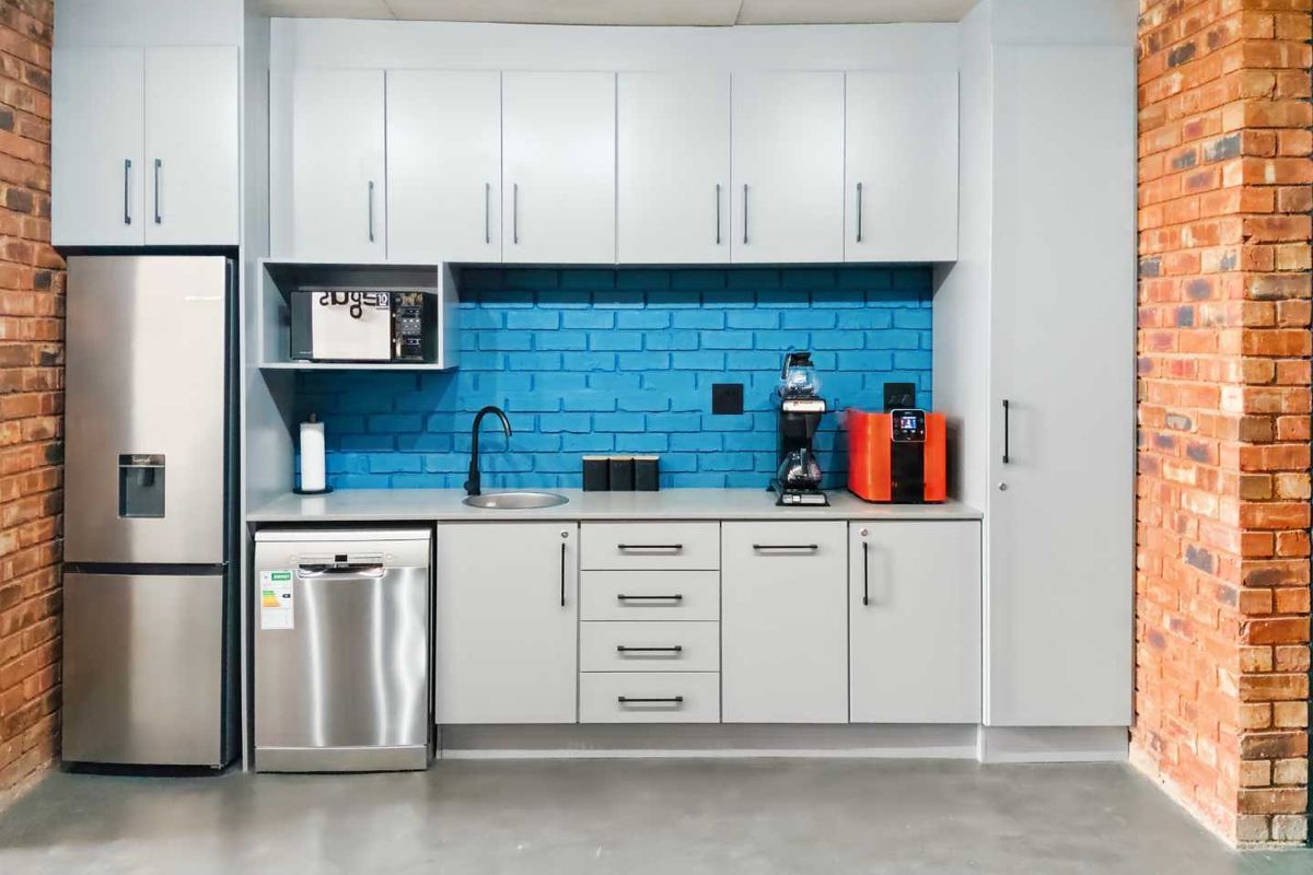 A modern office kitchenette featuring grey cabinetry, a blue brick backsplash, stainless steel dishwasher, and exposed brick side walls.