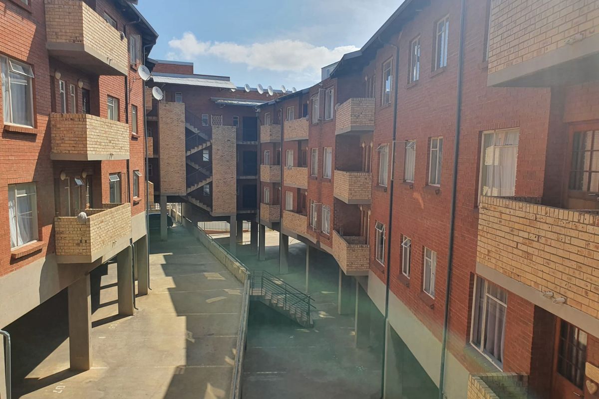 Exterior view of a multi-story apartment complex featuring red brick facades, private balconies, and a central paved courtyard area with concrete structural supports.